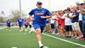 Mac McWilliams sprinting off the practice field, stopwatch in hand, while fans hold up two-for-one snack coupons.