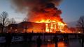 Charred regional parliament facade smoldering at dusk, firefighters silhouetted, hoses arcing, crowds watching behind barricades, banners drooping like wilted sentences.