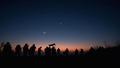Thin crescent moon sidling up to a bright star near Virgo, twilight gradient sky, silhouettes of bewildered backyard astronomers clutching coffee like ceremonial offerings.