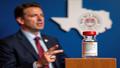 Texas health official gestures toward a map while a badge-shaped vaccine vial glints on the podium.