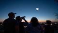 A family of stargazers squinting upward as a dim, unseeable silhouette hangs over a suburban skyline.