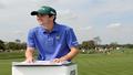 Keegan Bradley at a podium on a tee box, holding a binder labeled “Vibes Only,” Bethpage Black looming like a stern gym teacher.