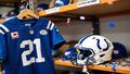 A Colts equipment manager placing a jersey on a shelf labeled Reserve/Retired as a fresh safety helmet is unboxed nearby, confetti hovering between sincerity and sarcasm.