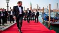 A tuxedoed guest stepping from a gondola onto a red carpet raft, photographers screaming like seagulls.