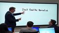 Microsoft's AI chief gestures toward a whiteboard labeled 'Don't Feed the Narrative,' as laptops nod in silent binary approval.