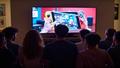 Crowd of fans refreshing screens as a mock Switch 2 preorder countdown blinks on a living-room TV.