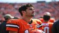 Flacco in Browns gear, helmet under arm, surveying the defense as cameras flash and fans blink in disbelief.