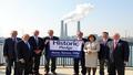 Officials pose under a banner reading ‘Historic Pledge’ as distant smokestacks clap politely, exhaling a light applause of haze.
