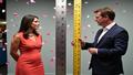 Woman stands confident, surgical scars visible, flanked by absurdly oversized rulers and confetti, while a news anchor compares compassion to inches on a chromed tape measure.