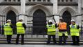 Conservators in high-vis vests surround a Banksy mural on the Royal Courts of Justice, measuring the wall like it’s a suspect in a lineup.
